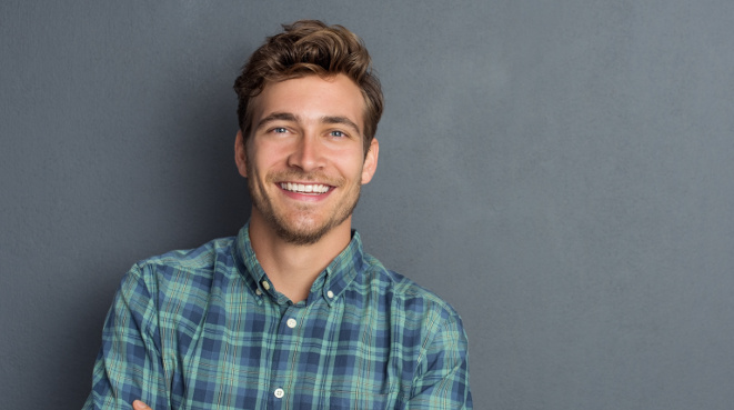 Young handsome man leaning against grey wall with arms crossed. Cheerful man laughing and looking at camera with a big grin. Portrait of a happy young man standing with crossed arms over grey background.